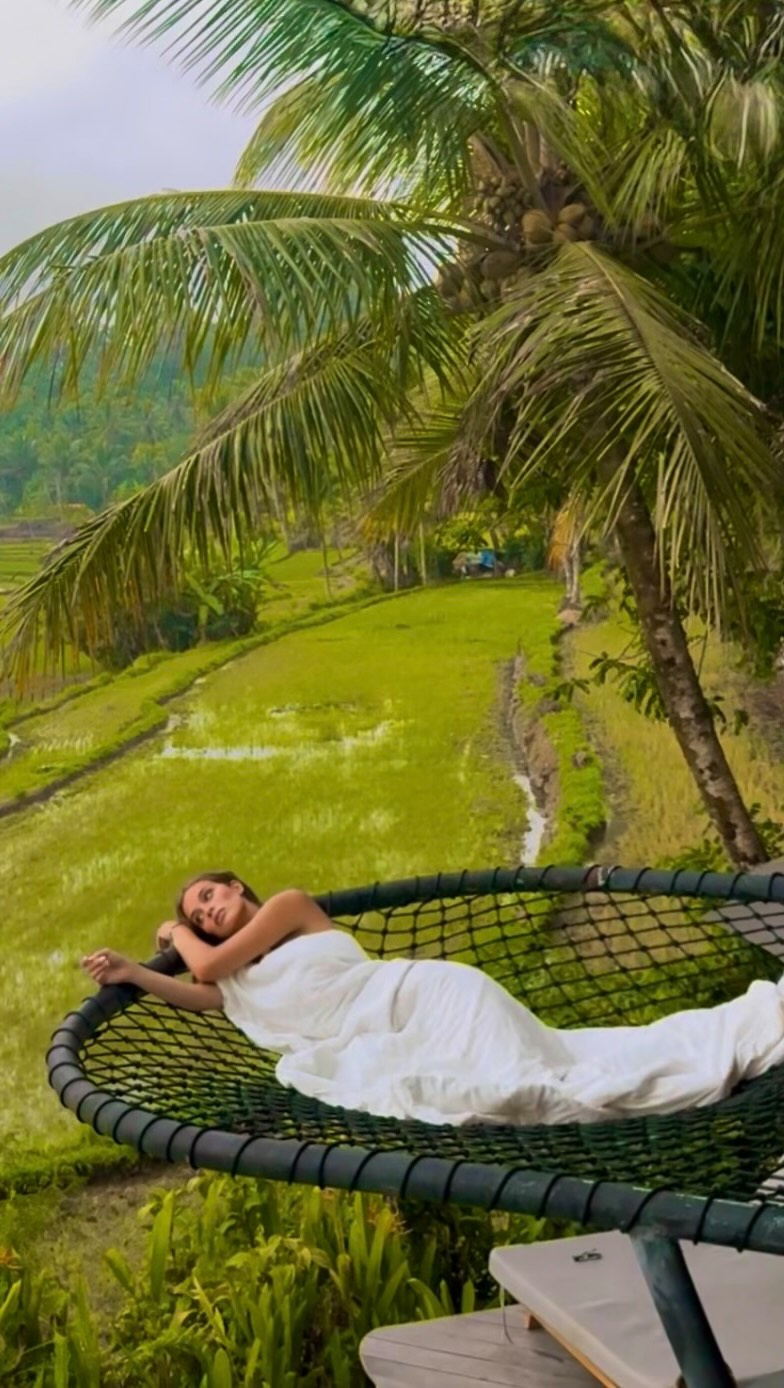Woman in a white dress lounging on a circular net hammock under palm trees in a lush tropical field setting.