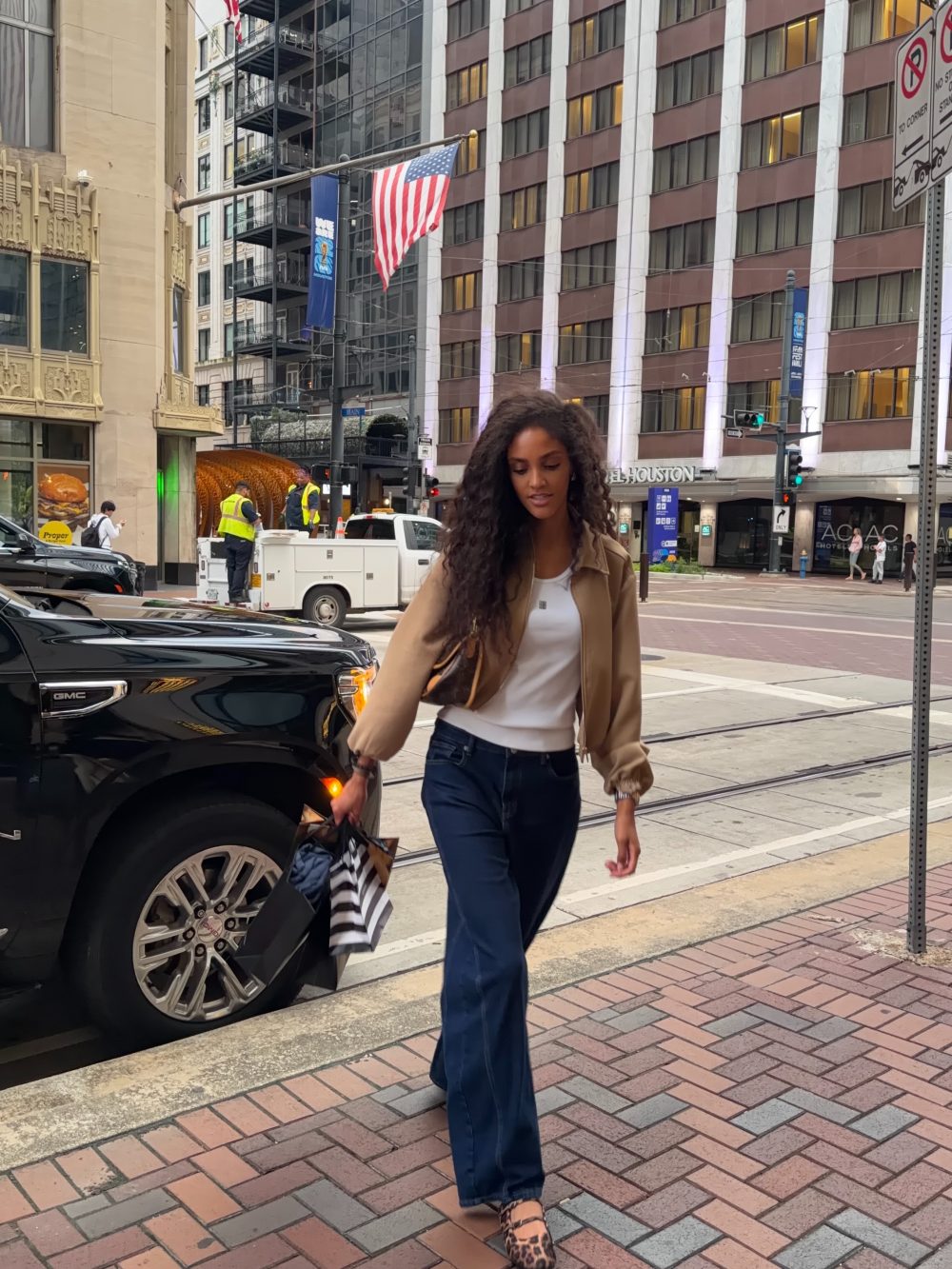 Woman in a beige jacket and blue jeans walks across a city street, carrying a striped bag; a black GMC truck and construction workers are in the background with a US flag overhead.