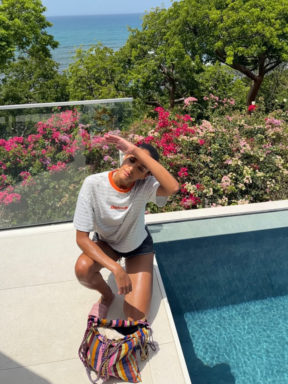 Person sitting by a blue pool with vibrant pink flowers and green trees, shielding eyes from the sun as the ocean is visible in the background.