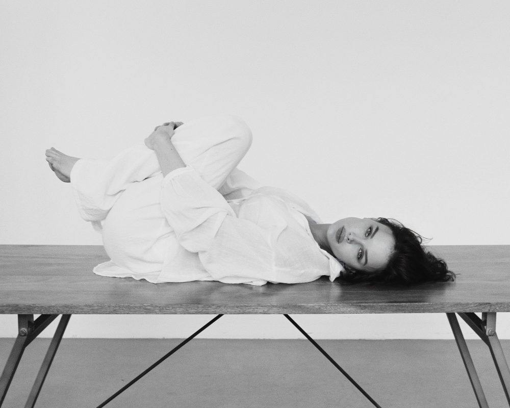 Person in a white robe lies on their side across a wooden table, head near the edge, looking toward the camera (black-and-white photo).