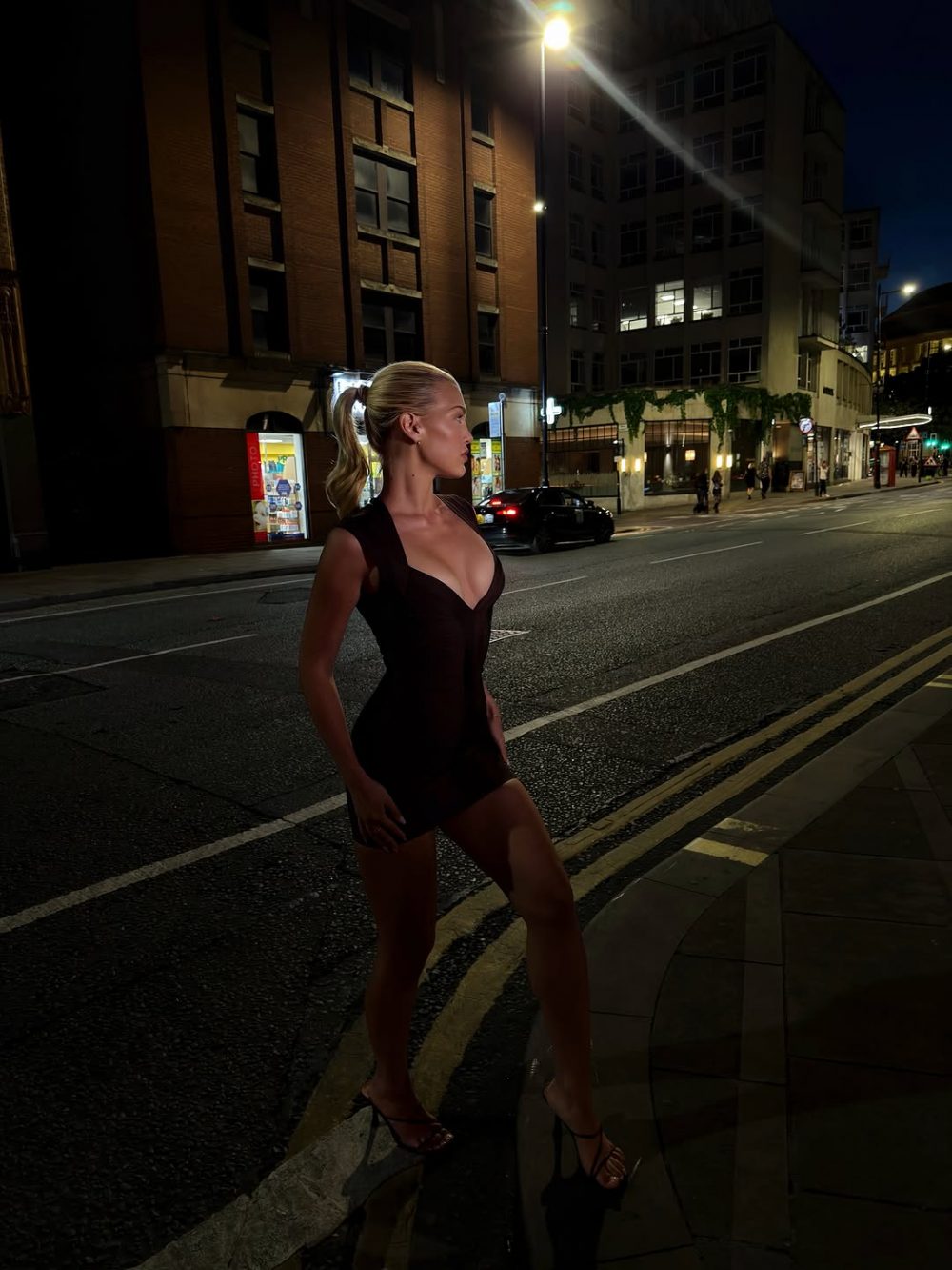 Woman in a black sleeveless dress and high heels posing on a city sidewalk at night, with streetlights and buildings in the background.