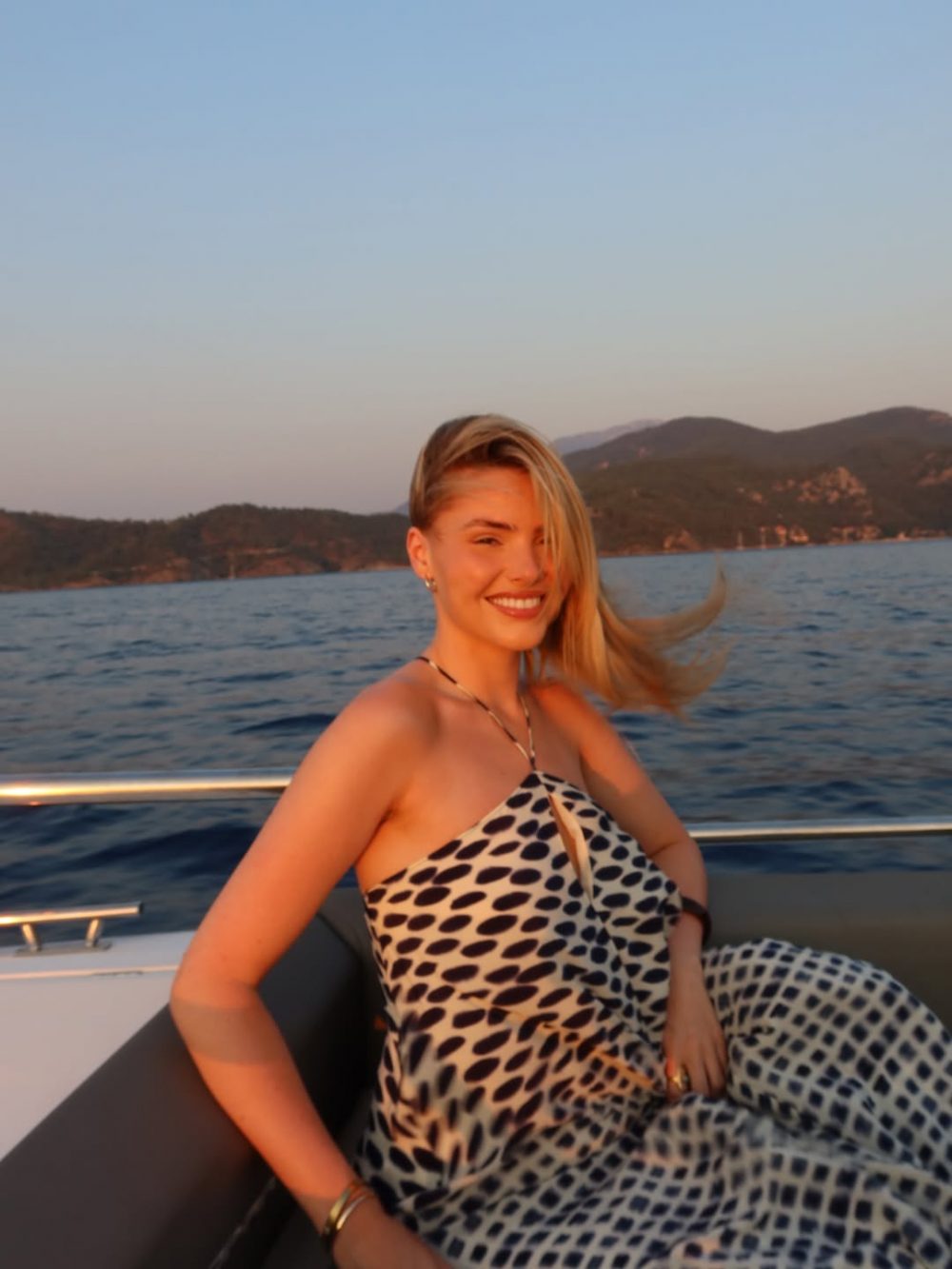 Woman smiling while seated on a boat at sunset, with calm sea and distant hills in the background.