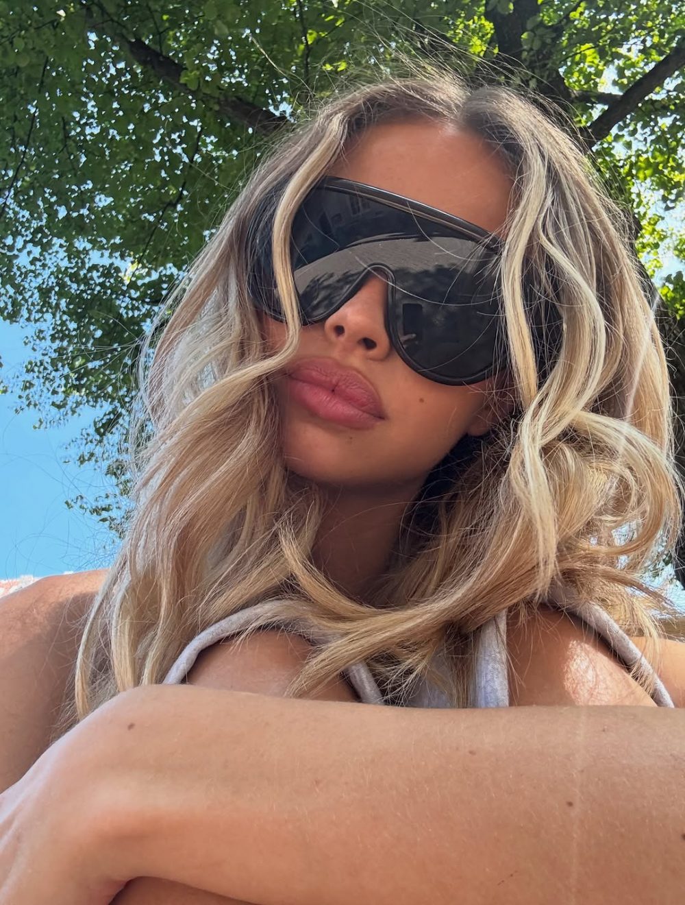 Selfie of a woman with long blonde hair wearing oversized dark sunglasses outdoors under a leafy tree.