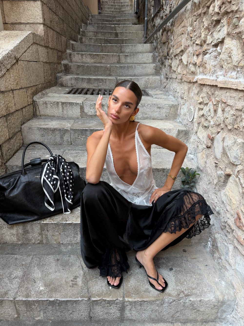 Fashionable woman poses on stone outdoor steps in a narrow historic alley, wearing a white lace top and black skirt with a thoughtful hand pose.
