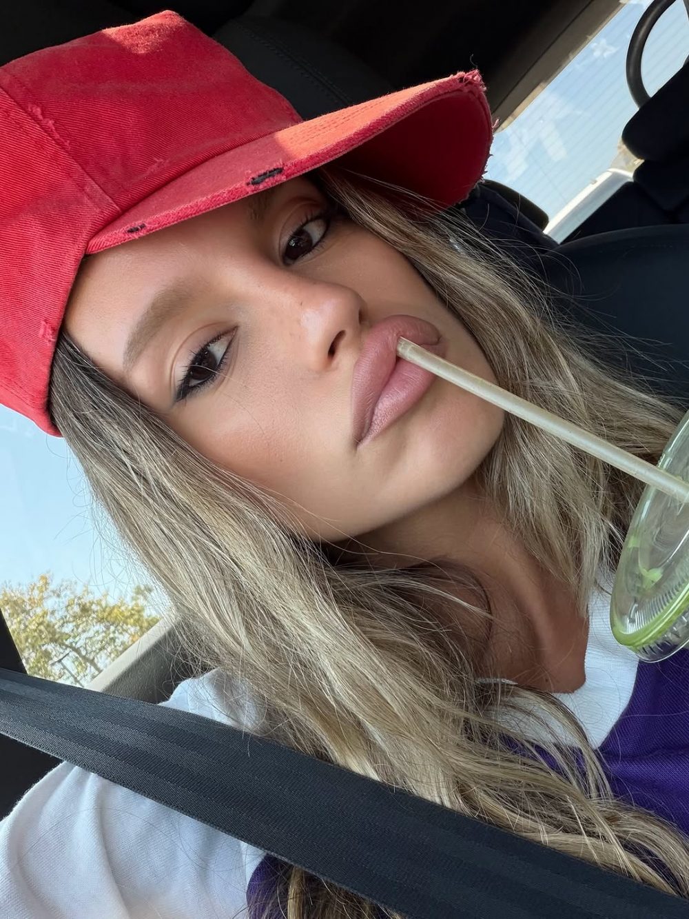 Young woman wearing a red bucket hat sits in a car with a seatbelt on, sipping from a straw in a green beverage cup.