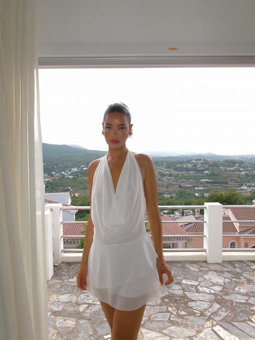 Woman in a white halter dress standing on a stone balcony with a hillside town in the background.