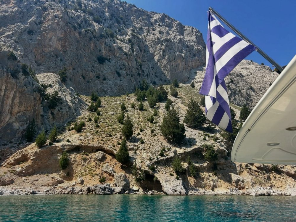 Flagged boat with blue-and-white striped flag against a rocky coastal cliff and clear turquoise water in the foreground.