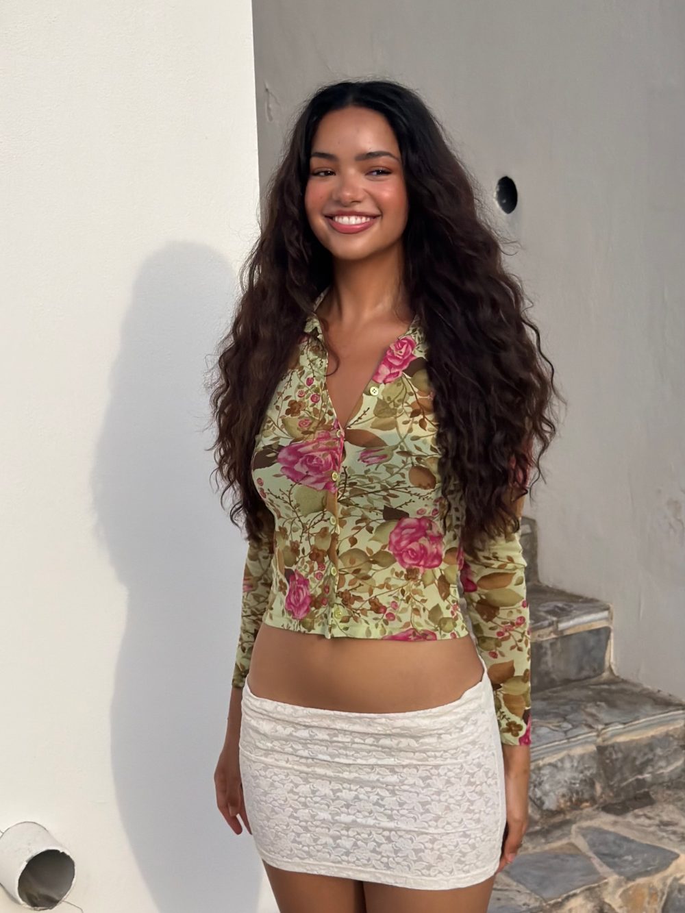 Smiling woman wearing a yellow floral button-down top and a white lace mini skirt, standing against a white wall outside.