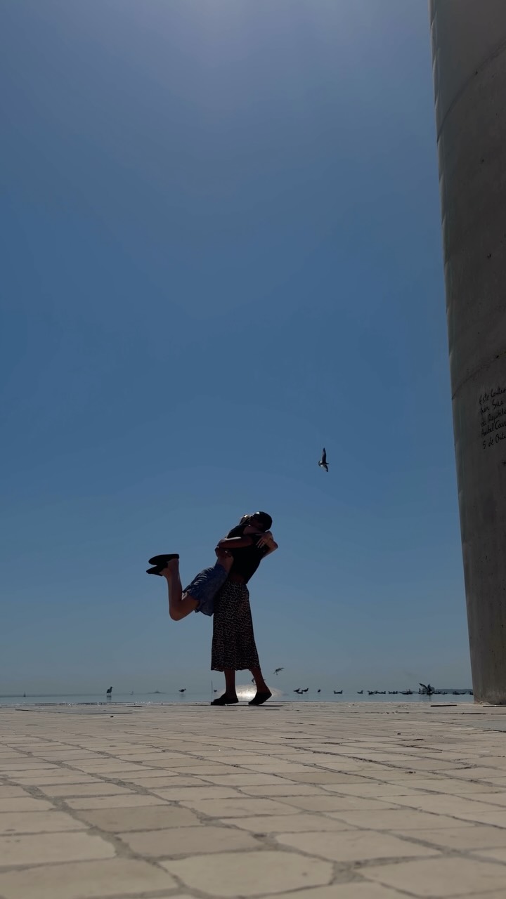 Couple embracing on a sunny seaside promenade, the man lifting the woman while blue sky and seagulls glare in the distance, with a tall pillar on the right.