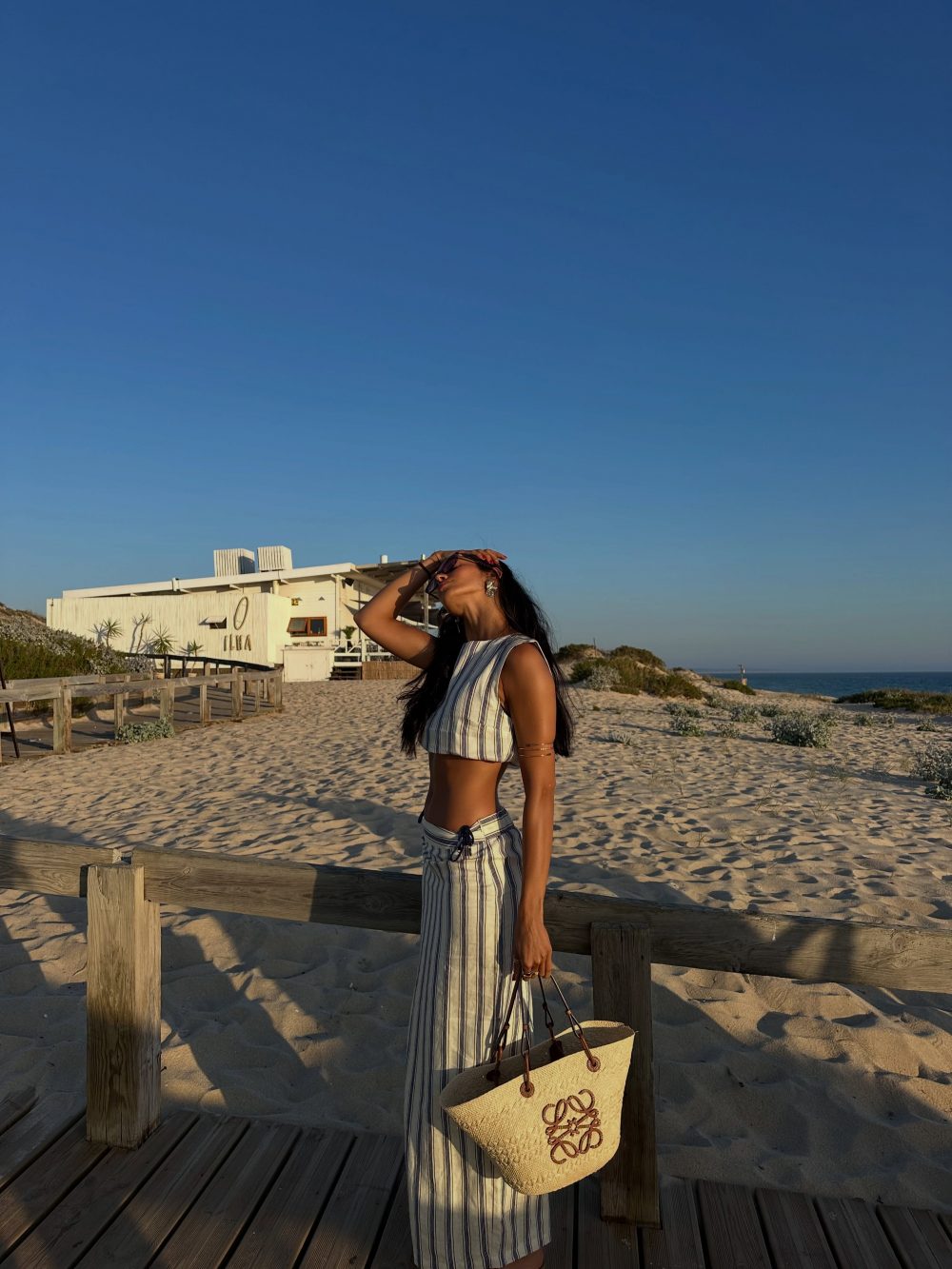 Woman in striped two-piece outfit on a sandy beach, carrying a straw bag near a wooden railing with a white building in the background under a clear blue sky