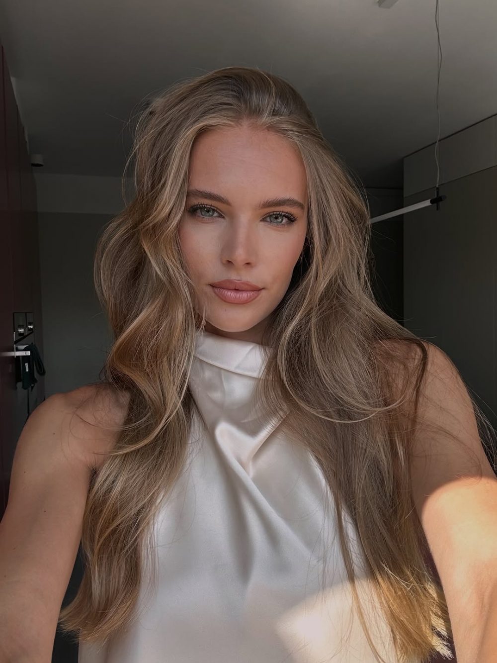 Portrait of a woman with long blonde hair in a white satin top, looking at the camera in a softly lit room.