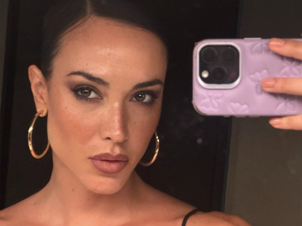 Close-up selfie of a woman with hoop earrings, holding a purple phone case, showing part of her face and makeup in a mirror selfie.