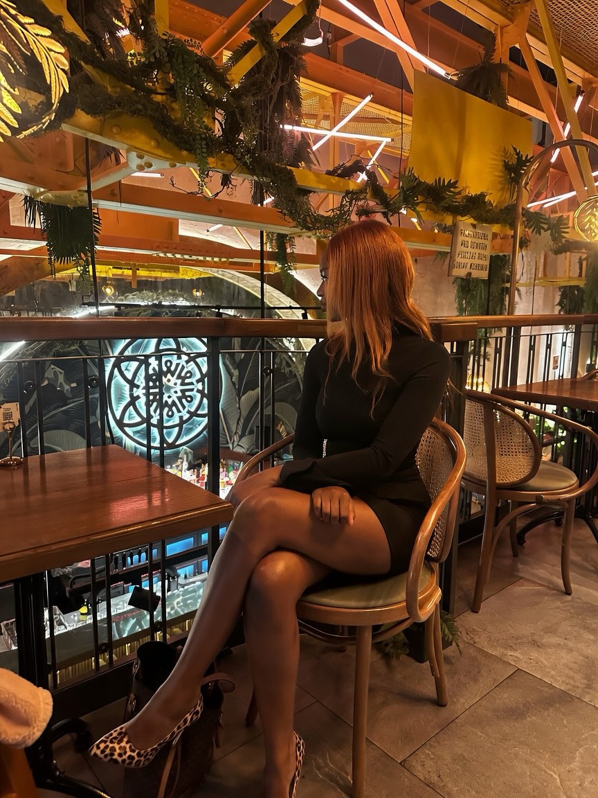Woman with red hair in a black dress sits on a wooden chair at a table in a plant-filled, warmly lit cafe, looking to her left.