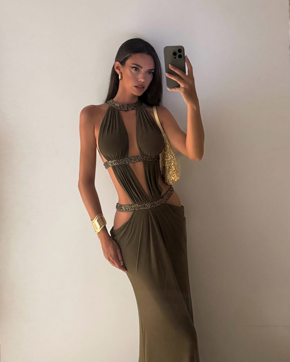 Woman in an olive green cutout gown taking a mirror selfie, with gold jewelry and a woven gold bag over her shoulder.