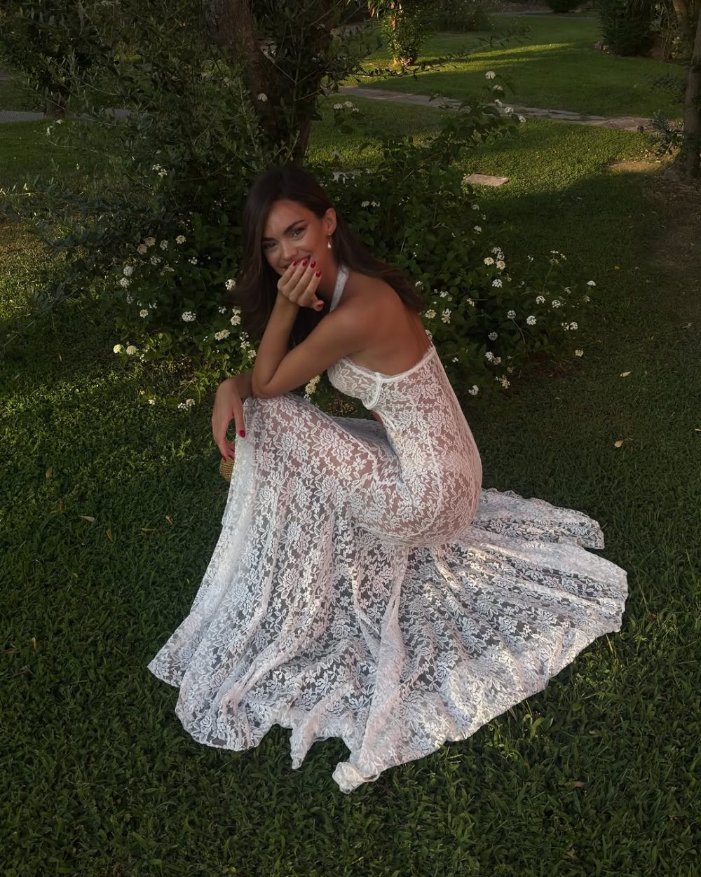 Woman in a white lace gown kneels on grass beside white-flowered bushes, smiling with her hand near her face and red nails.