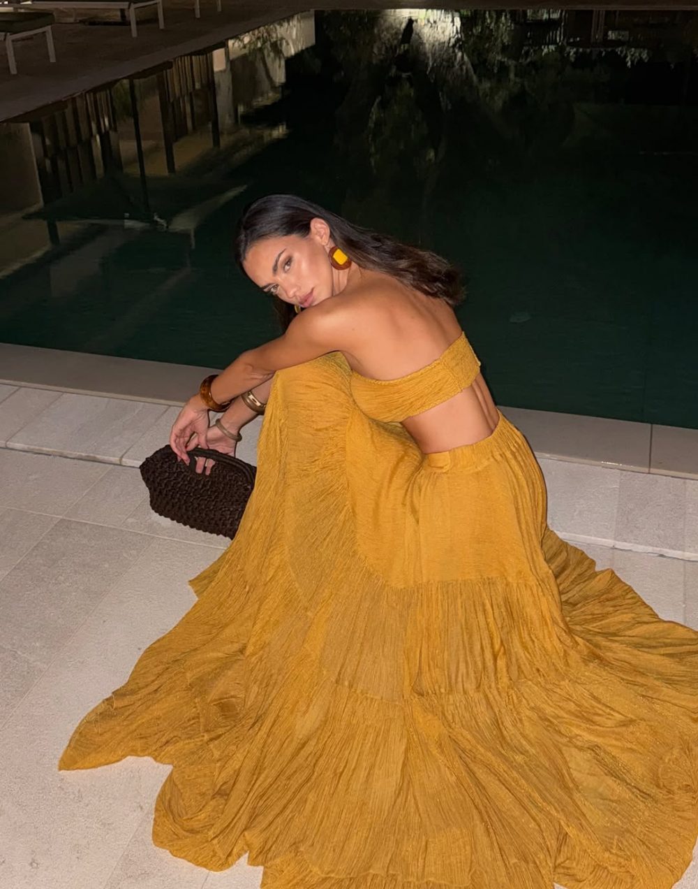 Woman in a mustard yellow off-shoulder top and long flowing skirt sits on a ledge by a dark pool at night, holding a woven brown clutch.
