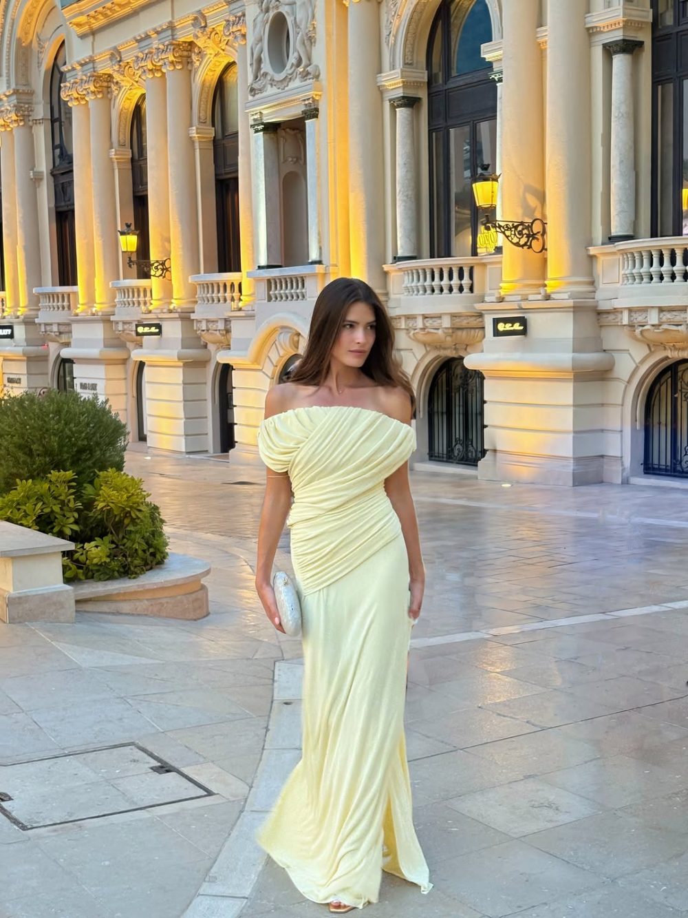 Woman in a pale yellow off-shoulder evening gown posing on a marble plaza in front of an ornate, illuminated building façade.