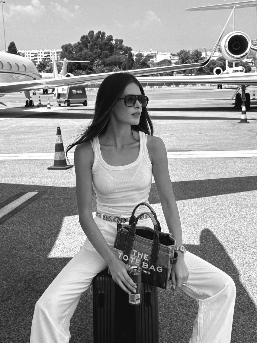 A woman sits on a black rolling suitcase on an airport tarmac, wearing a white tank top and white pants, sunglasses, and a tote bag; she holds a drink can and gazes to the side with a jet and cones in the background.