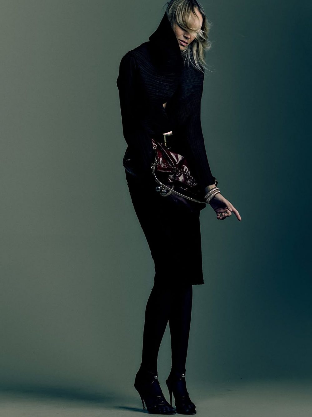 Fashion model in a black textured dress with a burgundy handbag, posing with one hand pointing downward, on a muted backdrop.