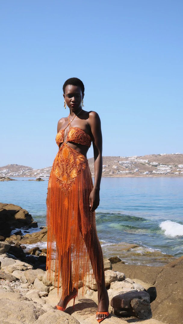 Woman in an orange fringe dress stands on a rocky beach with blue water and clear sky behind her.