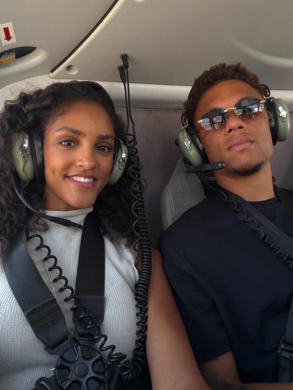 Two people wearing aviation headsets sit side by side in a helicopter, smiling at the camera.