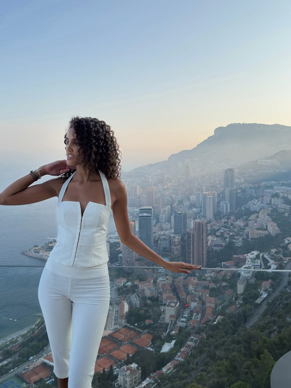 Woman in a white halter jumpsuit stands on a glass balcony, overlooking a coastal city and hills at sunset/sunrise behind her.