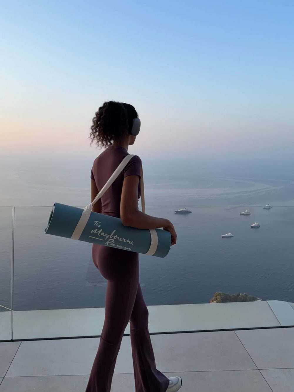 Woman with headphones carrying a rolled yoga mat gazing over a calm sea at sunset during golden hour.
