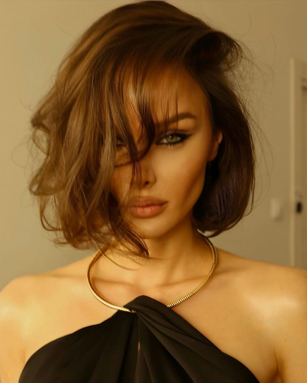 Portrait of a woman with a tousled brown bob, smoky eye makeup, and bare shoulders wearing a gold necklace.
