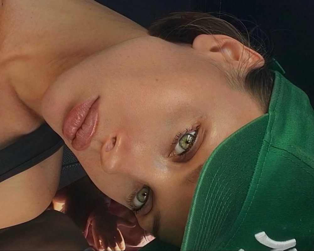 Close-up of a woman’s face resting on a surface, eyes open and looking toward the camera, wearing a green cap over her hair.