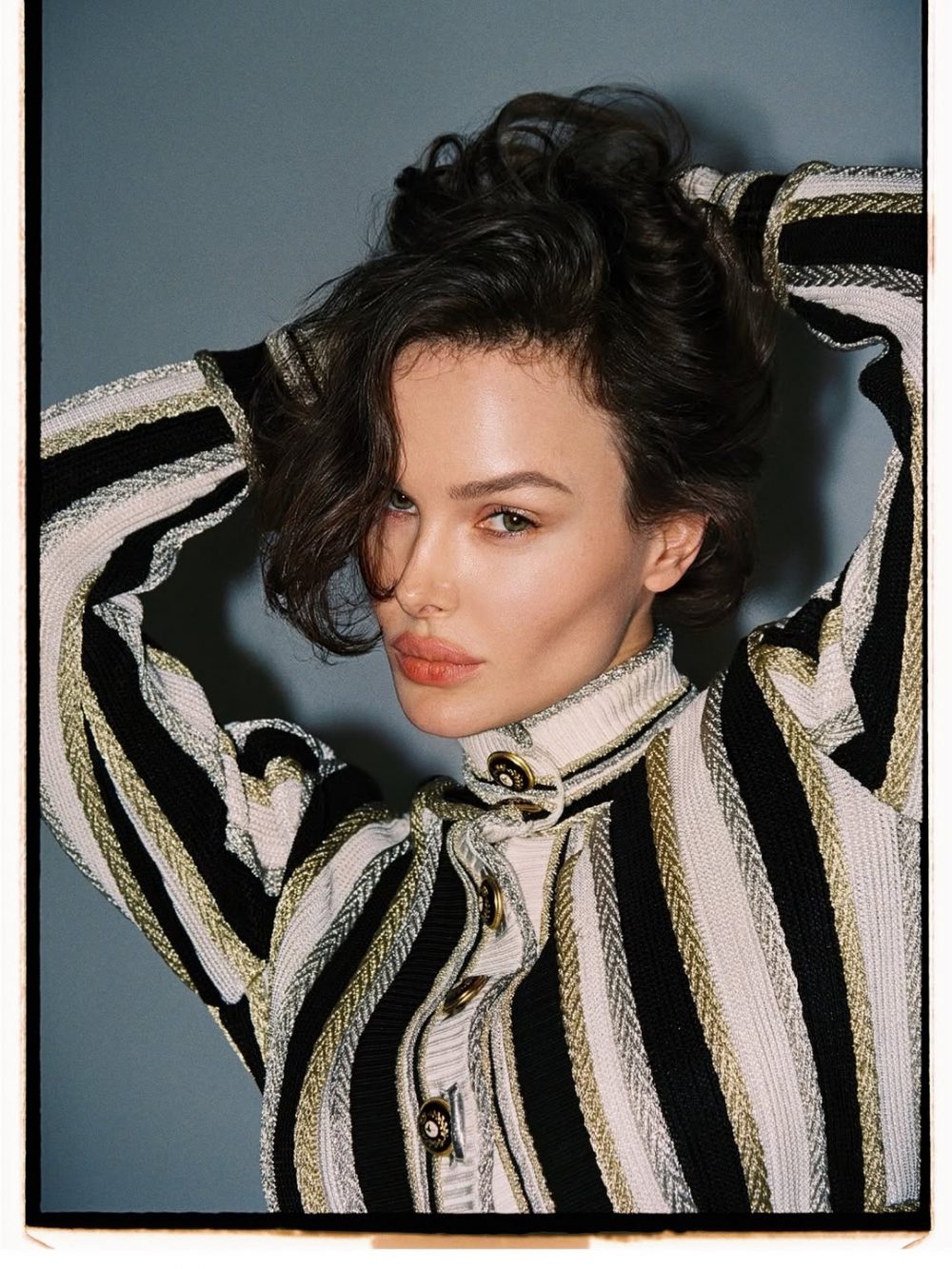 Portrait of a woman with short dark hair wearing a striped black, white, and gold knit sweater, hands in her hair.