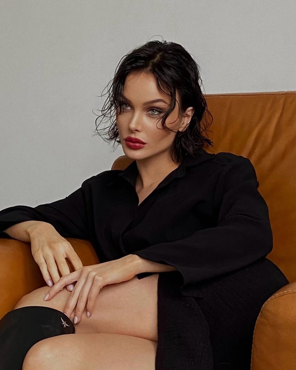 Woman with short black hair and red lipstick sits on a brown leather chair, wearing a black shirt and gazing at the camera.