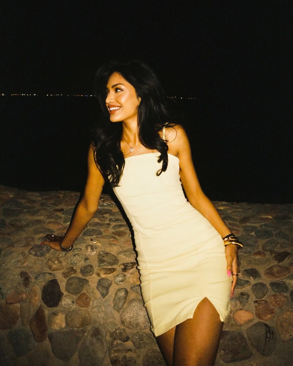 Woman in a white strapless mini dress smiles while leaning on a stone seawall at night, dark hair flowing down.