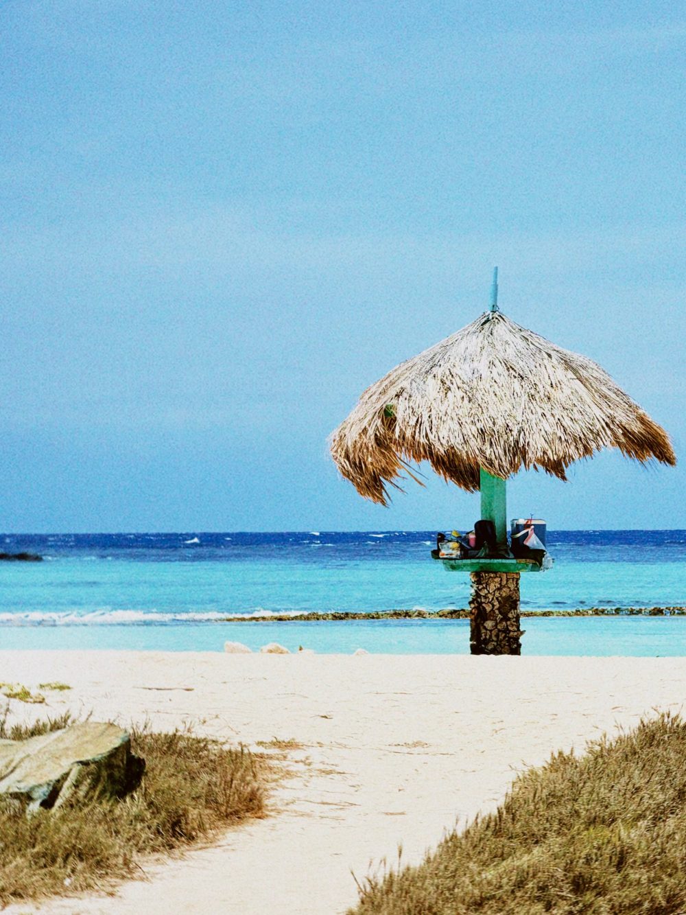 Thatched beach umbrella on a wooden post over clear turquoise water and white sandy beach under a blue sky.