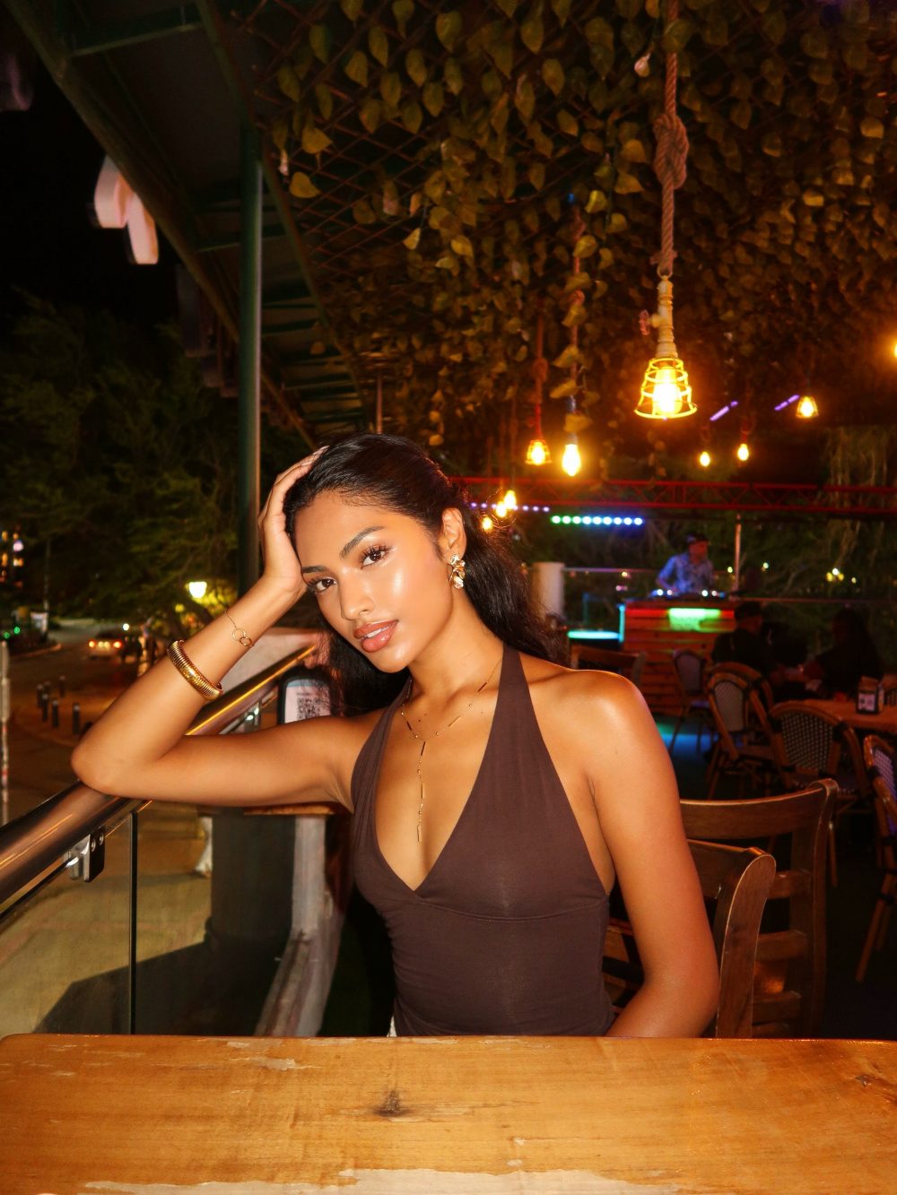 Woman leaning on a wooden railing at night, wearing a brown halter top and gold jewelry, with warm hanging lights in the background A glossy, sun-kissed look (informal setting).