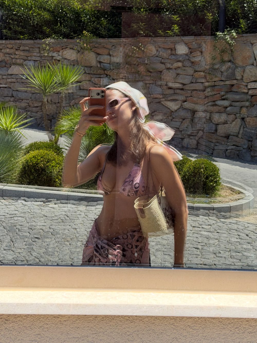 Woman in a pink bikini taking a selfie with a smartphone, reflected in a car window, with palm plants and a stone wall behind.