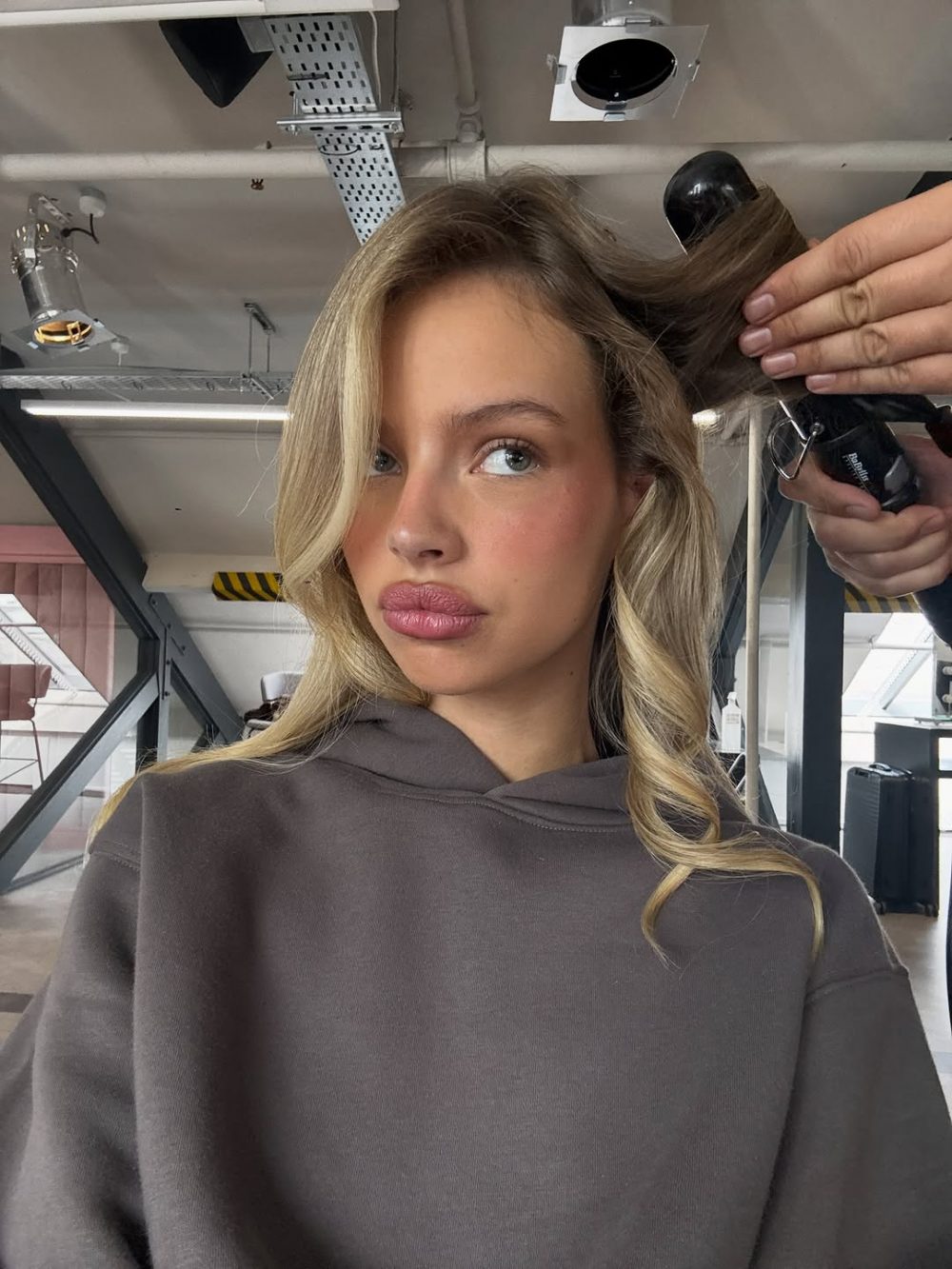 Young woman with blond hair in a gray hoodie sits in a modern salon as a stylist trims the top of her head with clippers.