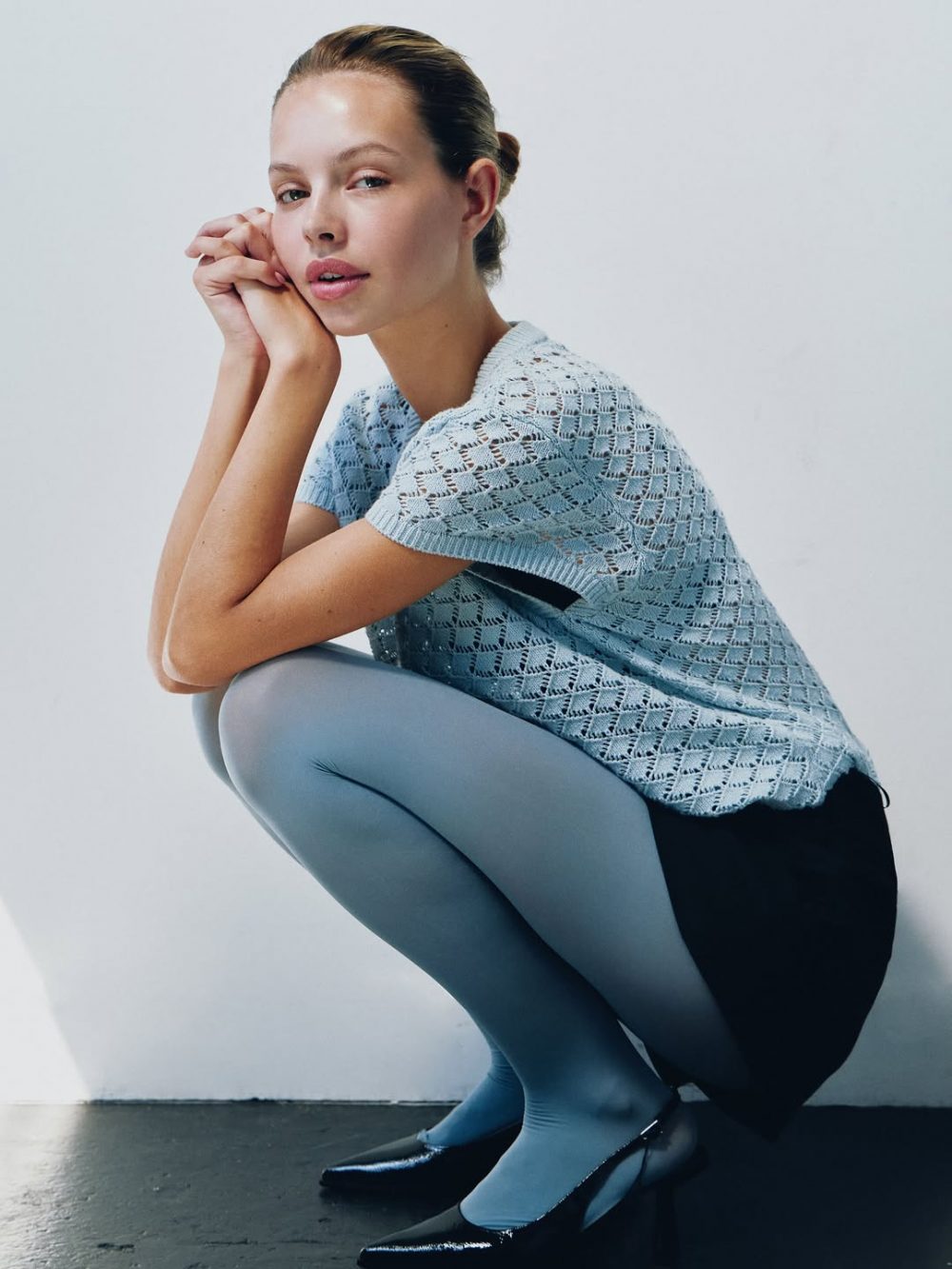 Woman crouching in a light blue knitted top and matching blue tights, hands resting near her face against a pale wall.