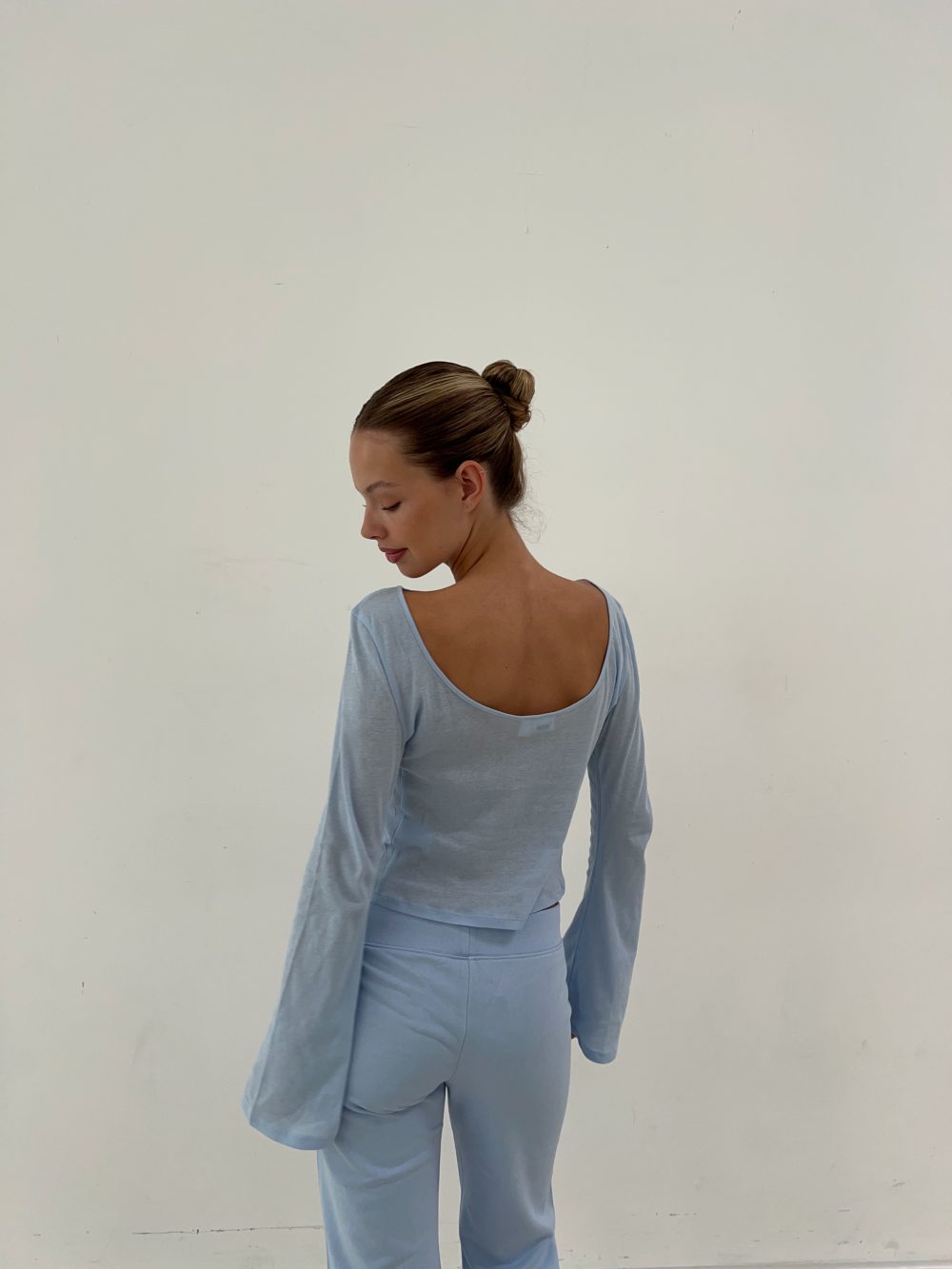Side view of a woman wearing a pale blue matching top and pants, hair in a bun, against a plain light wall.