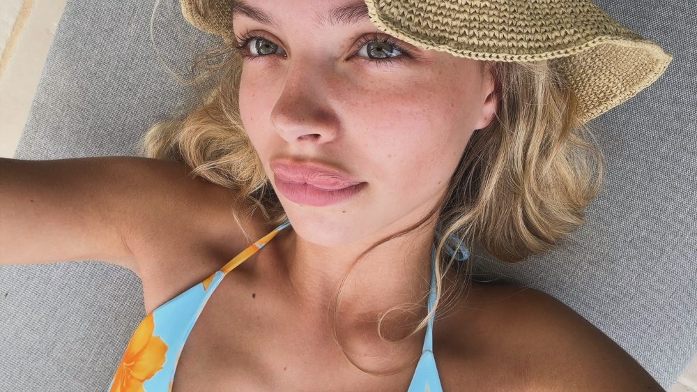 Young woman sunbathing in a woven straw hat, wearing a blue/orange swimsuit top, lying on a gray lounge surface.