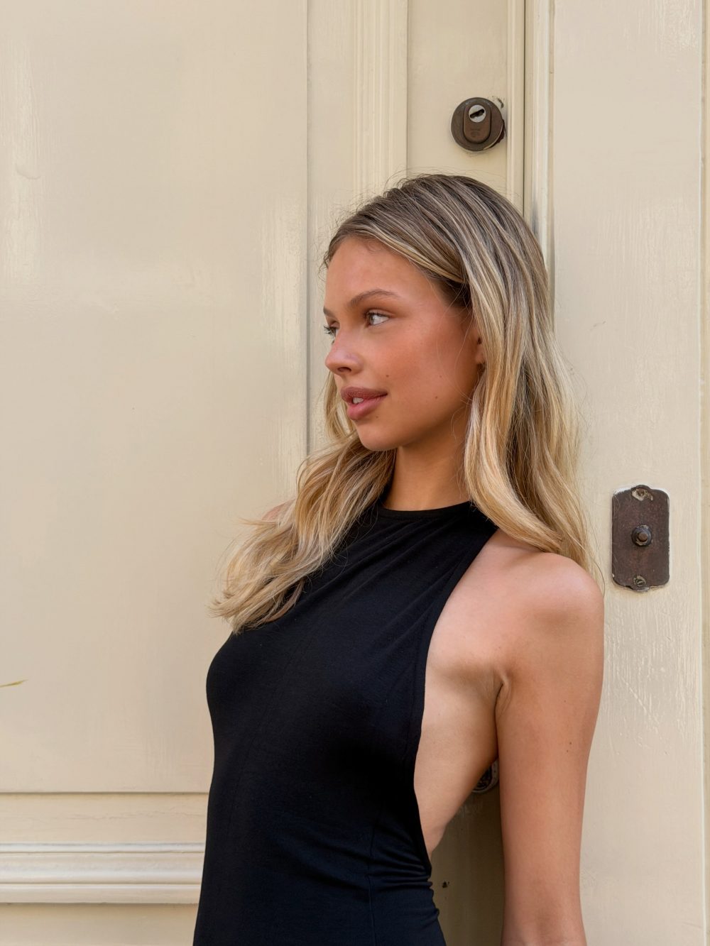 Side profile of a young woman with blonde hair wearing a black sleeveless top, standing against a pale beige door with a round lock visible behind her.