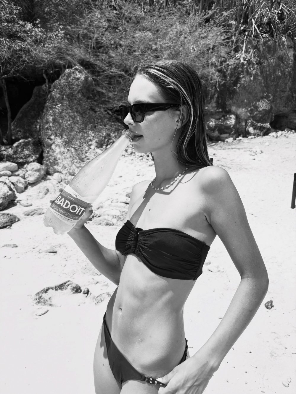 Woman in a black bikini with sunglasses drinks from a bottle on a sunny beach.