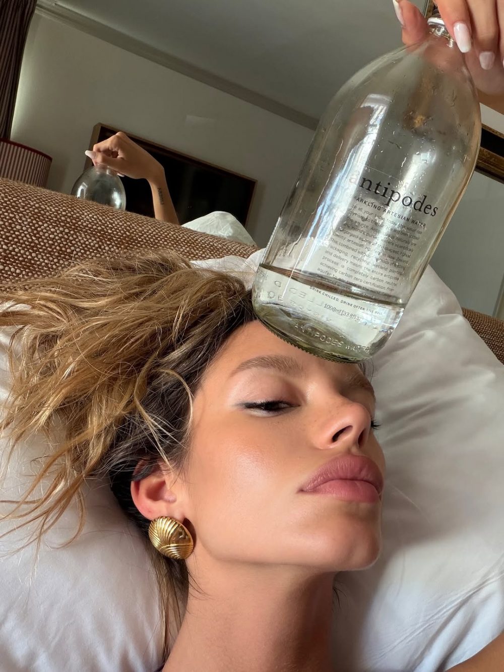 Woman lying on a bed with a large glass bottle balanced on her forehead as another hand holds it above her head.
