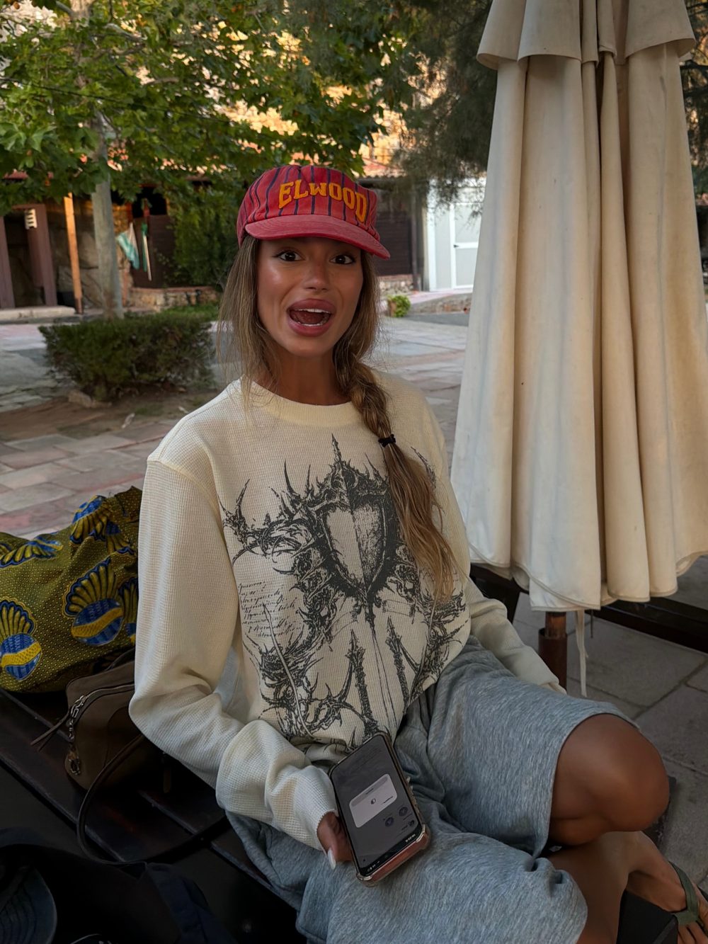 Woman in a red Elwood cap and beige sweater sits outdoors, smiling, while holding a smartphone. A large beige umbrella is nearby.