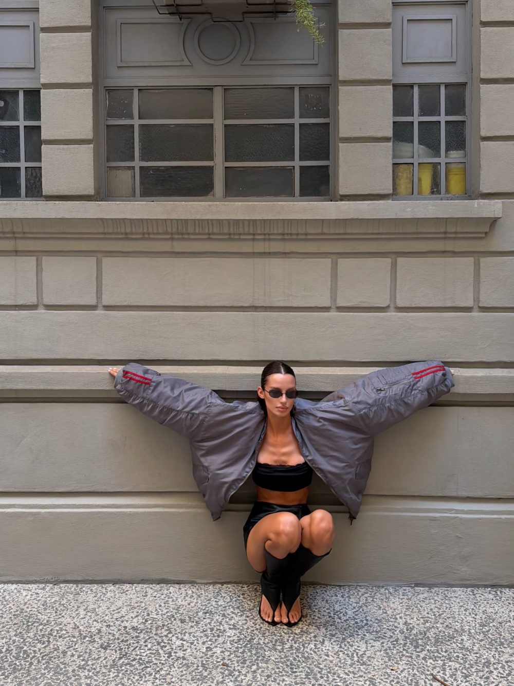 Woman wearing an oversized gray jacket with arms spread along a stone wall, sunglasses, seated on a sill/balustrade.
