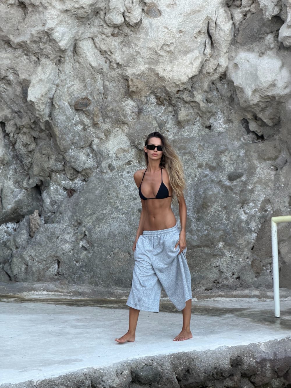 Woman in a black bikini top and gray wide-leg pants standing barefoot on a concrete surface against a rugged rock wall.