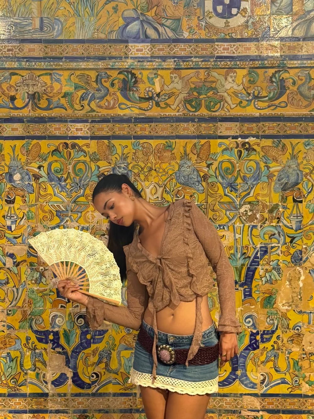 Woman posing in a sheer brown blouse and denim skirt, holding a lace fan in front of a vibrant tiled wall pattern.
