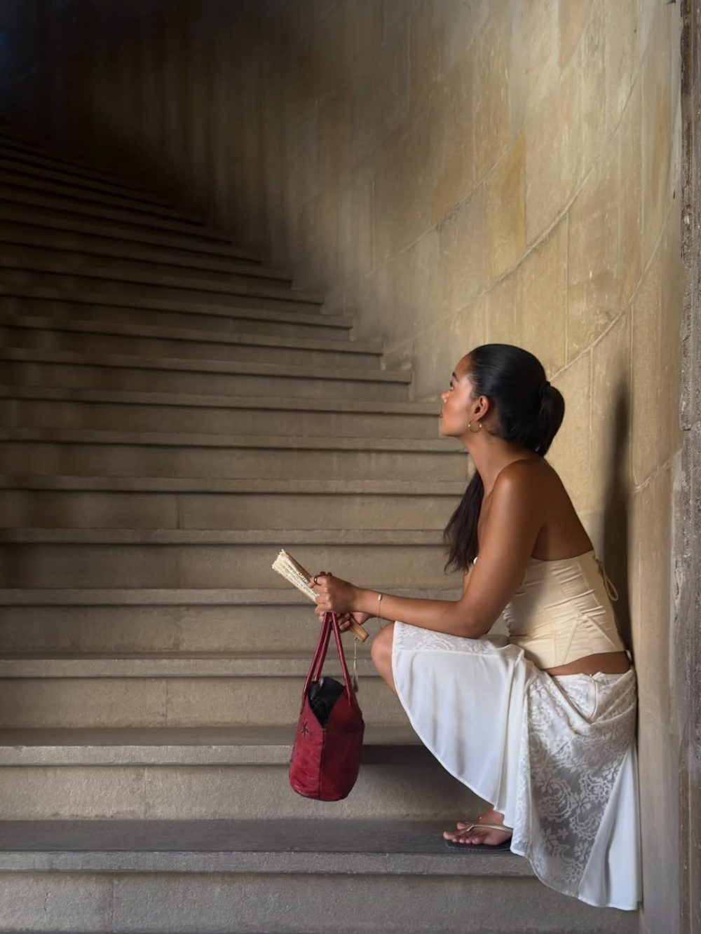 Woman in a cream corset top and white lace skirt sits on stone stairs, holding a fan and a red handbag, facing left.