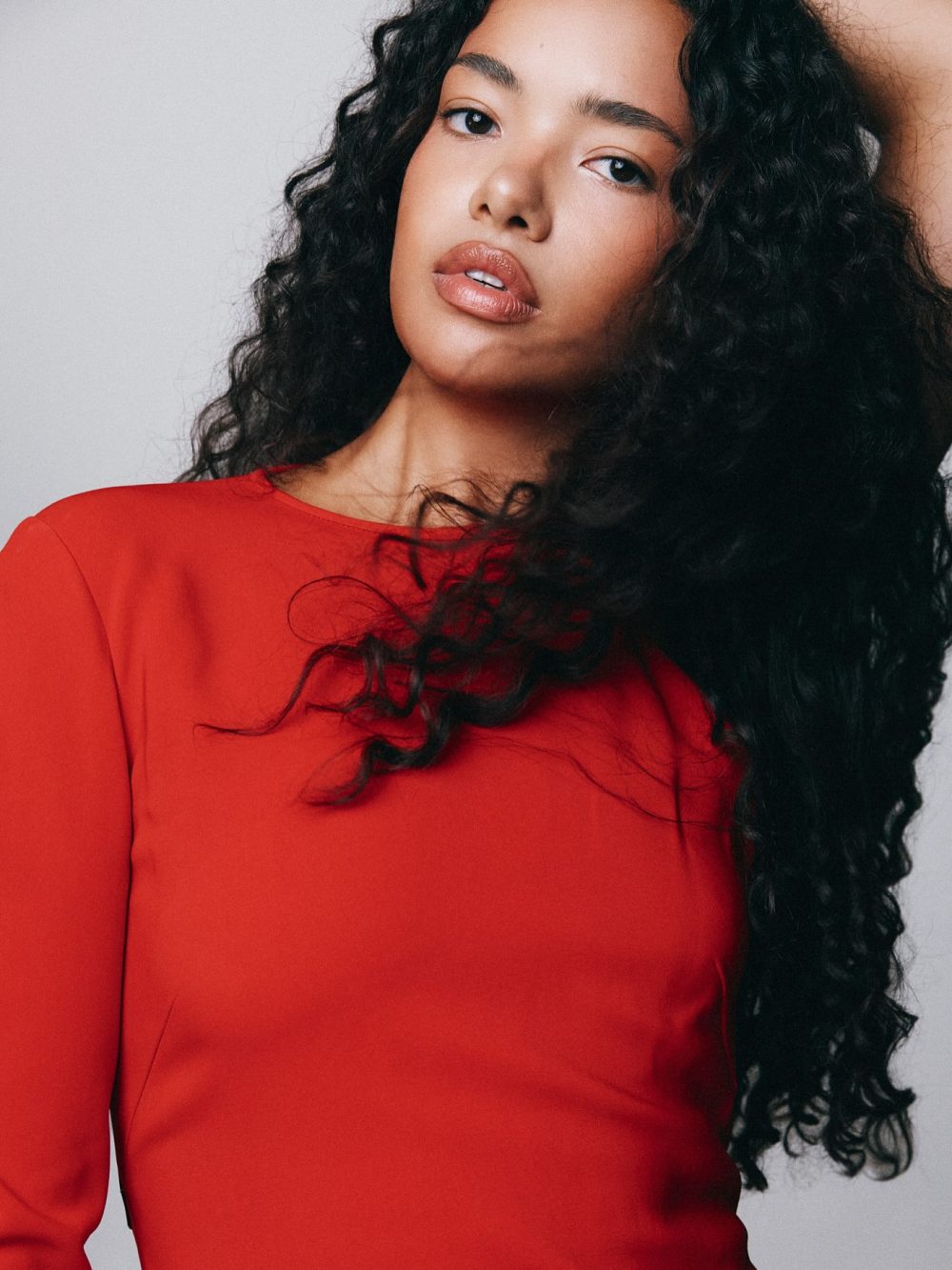 Portrait of a woman with long curly hair wearing a red top, posed against a light gray background, looking slightly to the side.