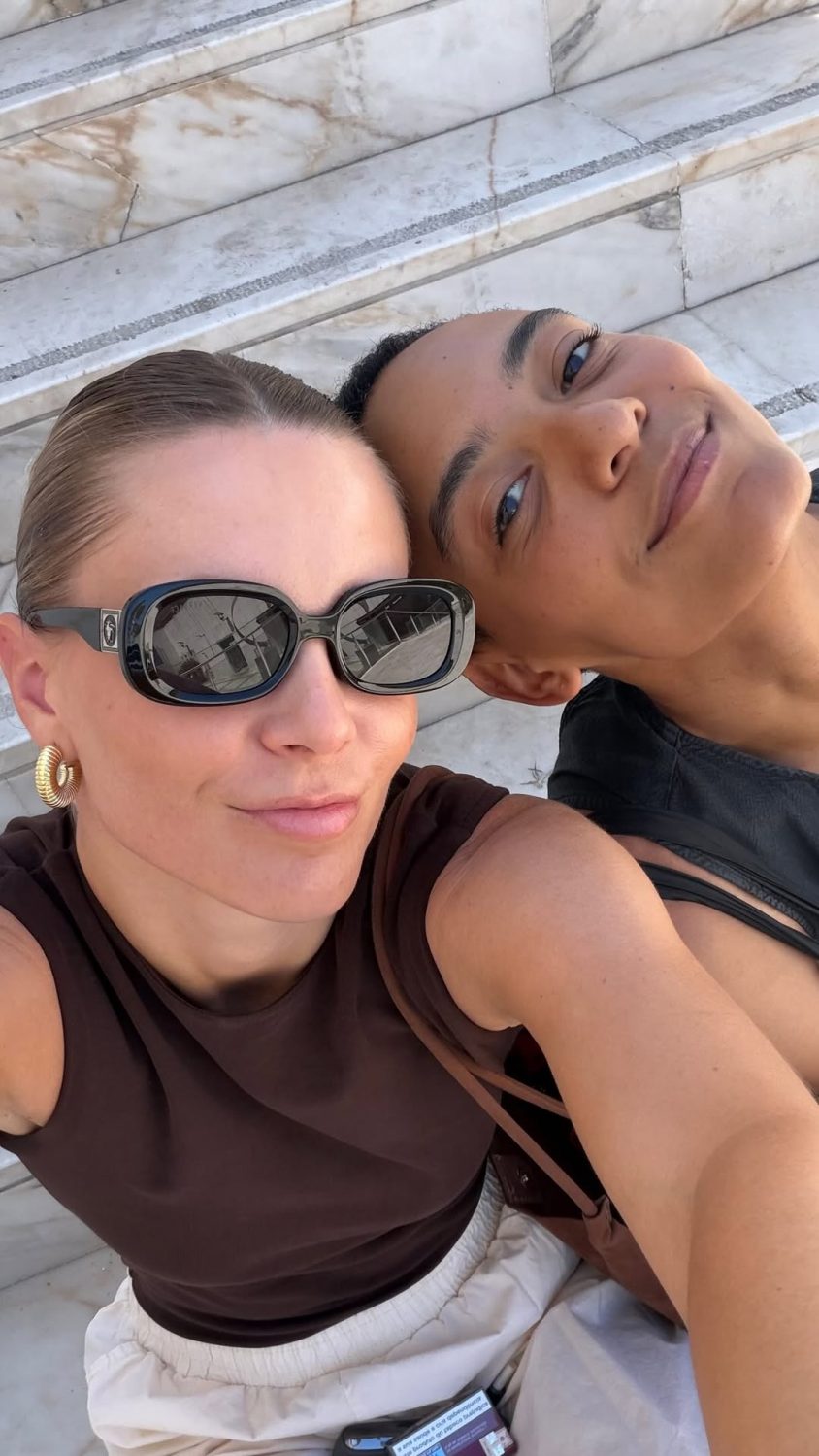 Two women pose for a selfie on marble stairs; one wears large sunglasses and gold hoop earrings.