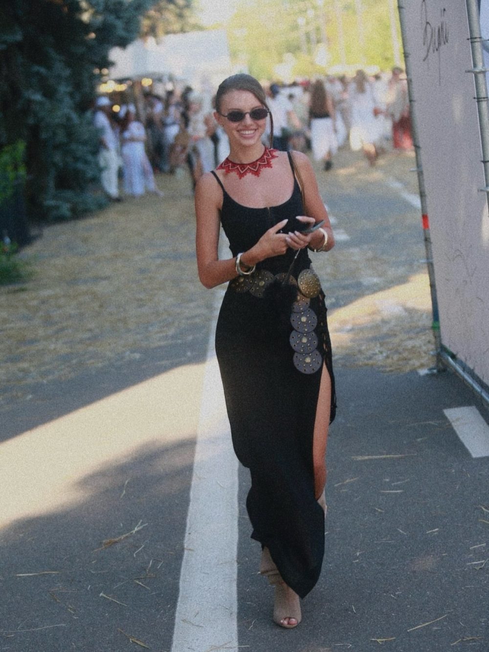 Woman in a black sleeveless dress with a red statement necklace, sunglasses, and a belt, walking on a sunny street while holding a phone.